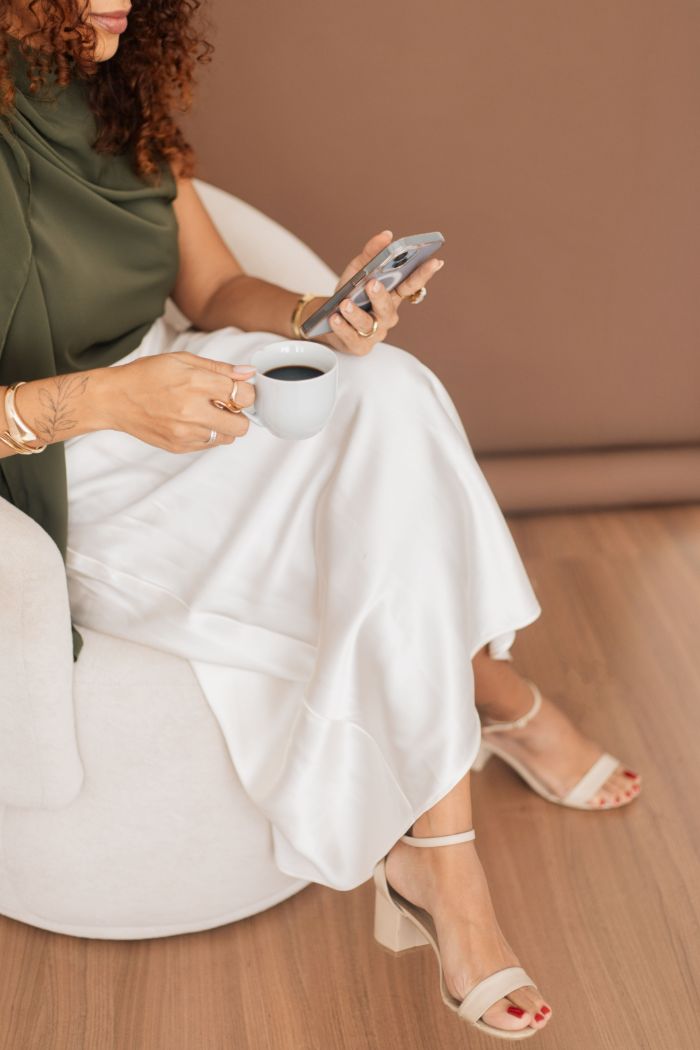 Xícara de café e celular no colo, sentada em uma poltrona clara