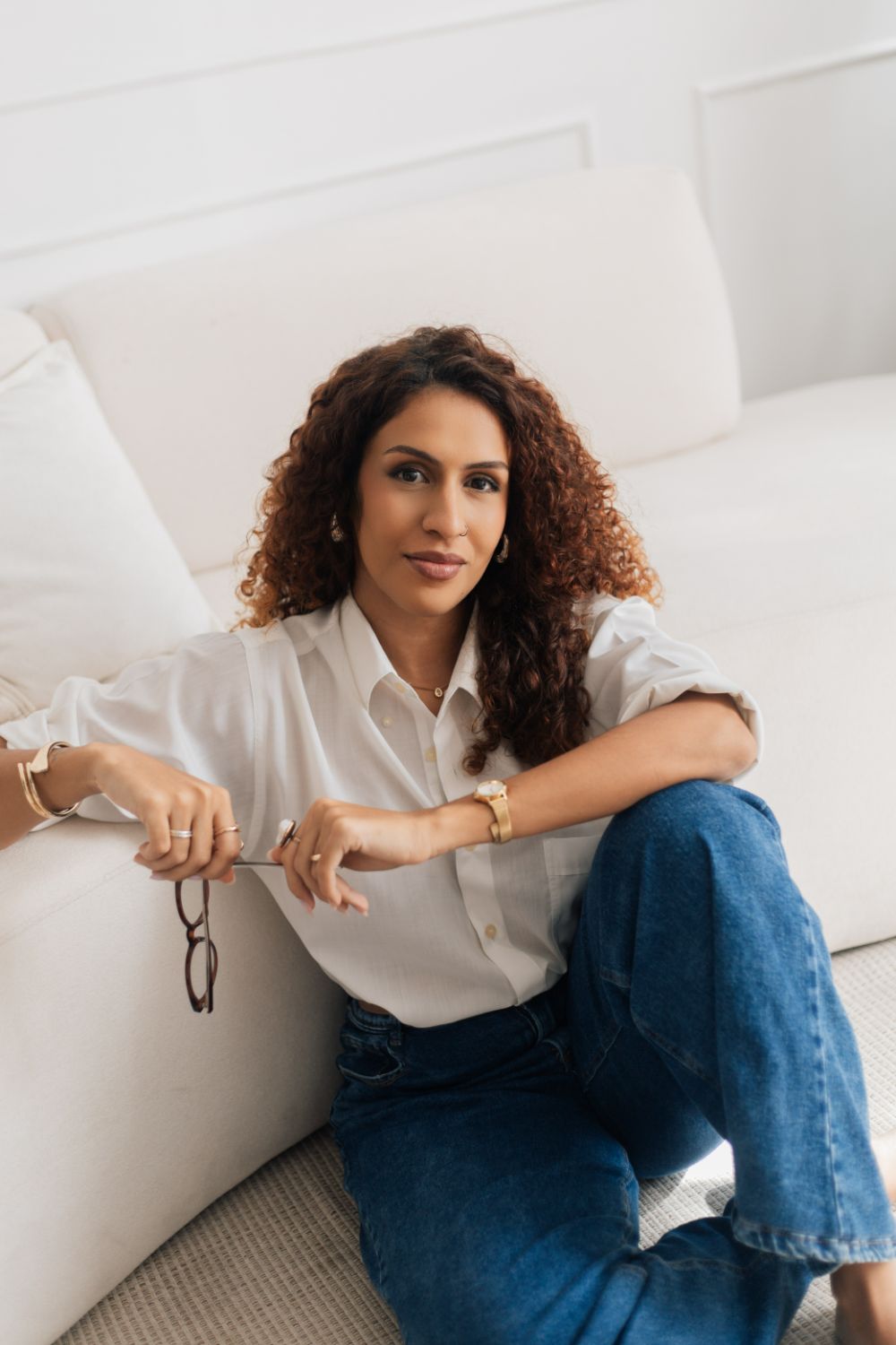 Aline Valente sentada no chão da sala, de camisa branca e jeans, sorrindo de leve